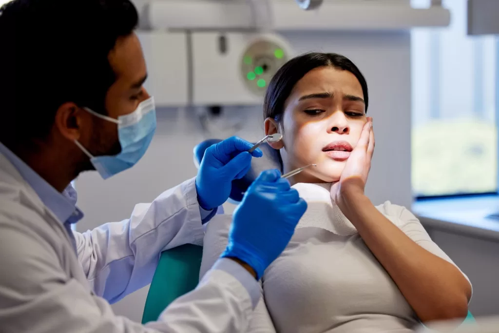 A dentist in a mask and gloves examines a patient holding their cheek, showing signs of discomfort after wisdom teeth removal.