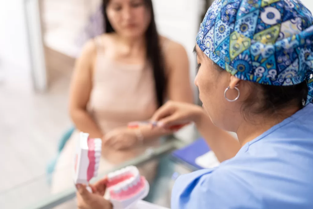 A dental professional in blue scrubs and a patterned cap explains oral care to a patient using a teeth model and toothbrush.