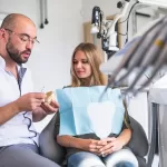 A dentist holding a model of teeth speaking with a patient holding a mirror.