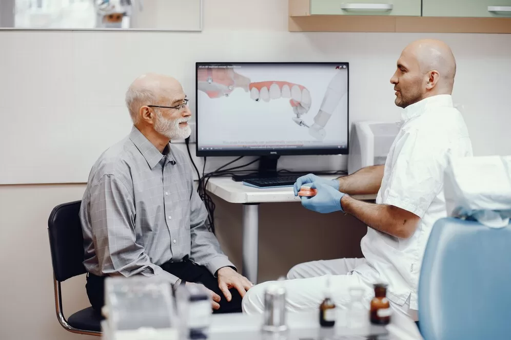 A dentist holding a model of teeth speaking with an elderly man in front of a screen showing teeth.