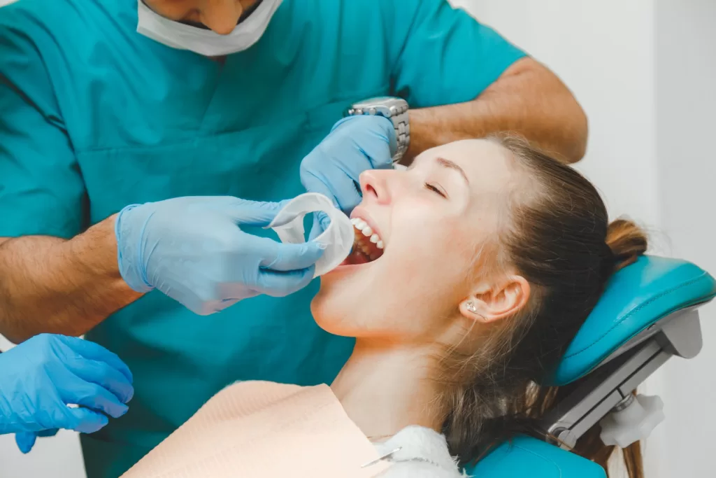 Dentist placing cotton roll in young female patient’s mouth during local anesthesia preparation at a dental clinic.