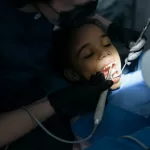 A child in a dentist's chair undergoes a procedure with the dentist using tools, highlighting a calm, professional setting.