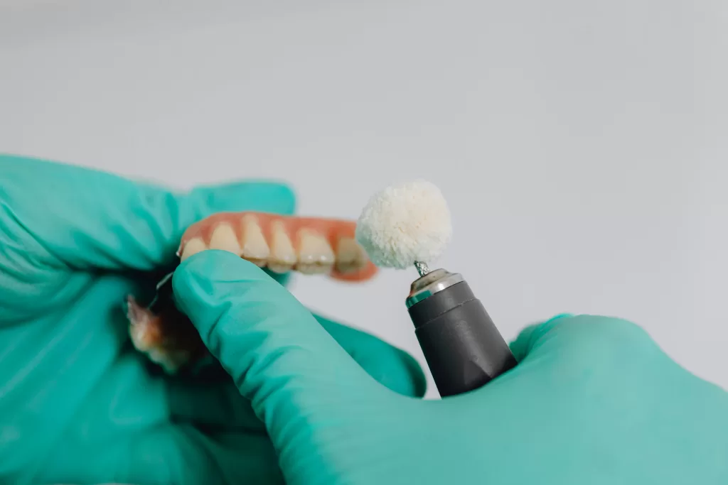 A dentist in blue gloves cleans dentures with a motorized brush, showing the plastic or ceramic material of the dentures.