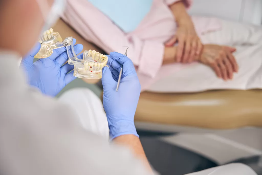 A dentist wearing blue gloves is holding a dental model to explain bone grafts to a seated patient.