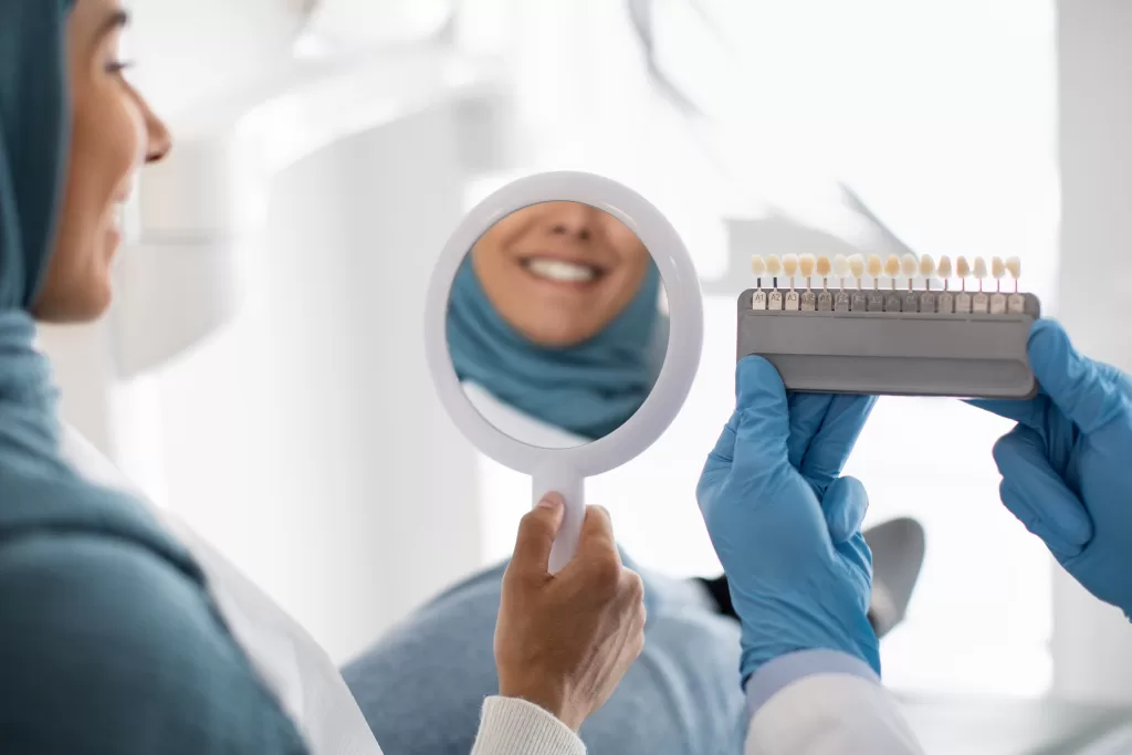 A patient smiles in a mirror as a dentist matches her tooth color for dental implants.






