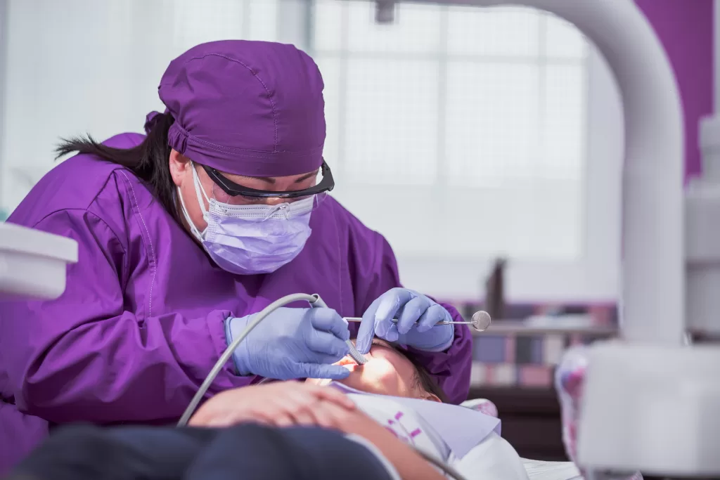 A dental professional dressed in purple scrubs, mask, and gloves performs an oral surgery procedure on a patient, using precision tools under bright clinical lighting.