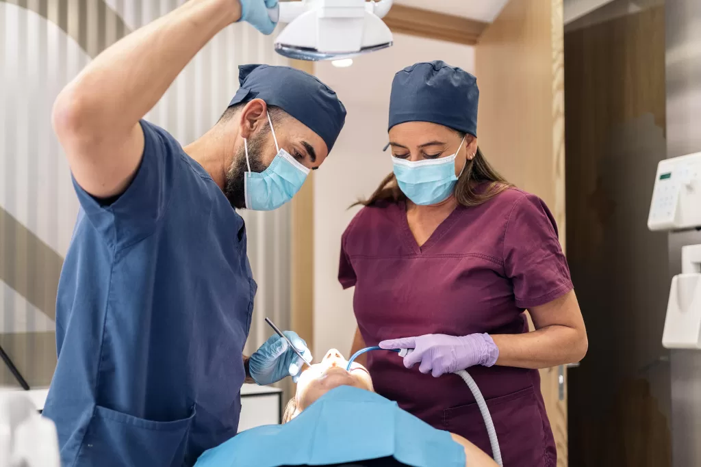 Two dental professionals wearing scrubs, masks, and gloves perform an oral surgery procedure under a bright overhead light, focusing on a patient reclined in a dental chair.
