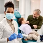 Female pediatric dentist wearing a face mask with a child patient and parent in a dental clinic.