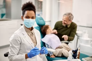 Female pediatric dentist wearing a face mask with a child patient and parent in a dental clinic.