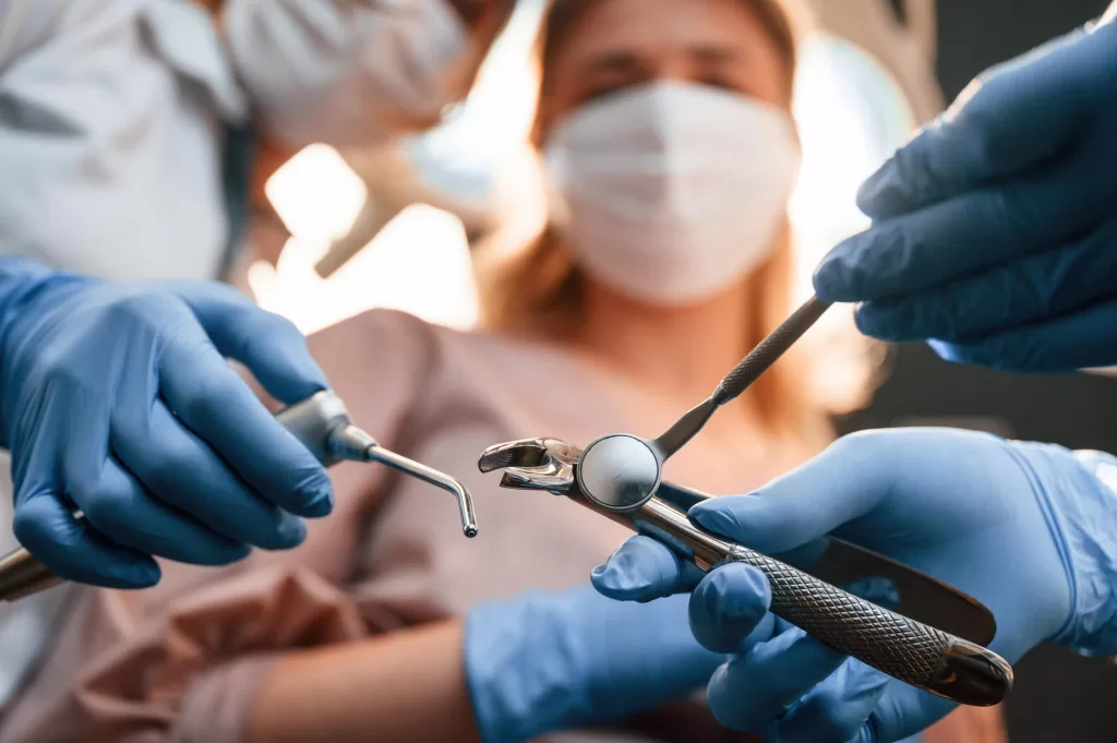 A dentist and assistant wearing gloves and masks prepare tools for a tooth extraction procedure, with a patient in the background.