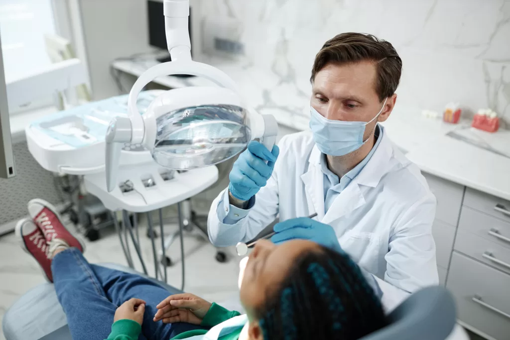 A dentist wearing a surgical mask and blue gloves performing an oral surgery procedure on a patient reclining in a dental chair, with a bright examination light focused on the patient's mouth.