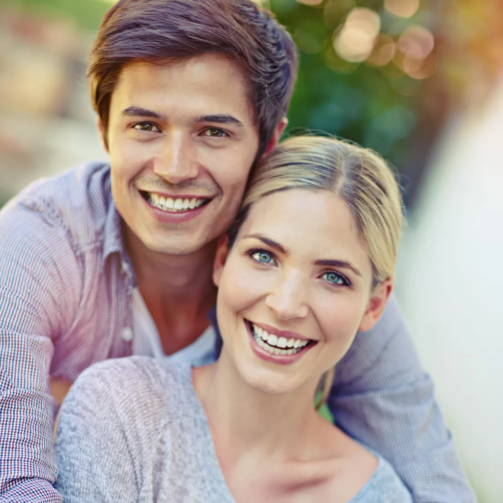 A smiling couple outdoors, with the man standing behind the woman and both looking directly at the camera, surrounded by soft natural lighting and a blurred green background.