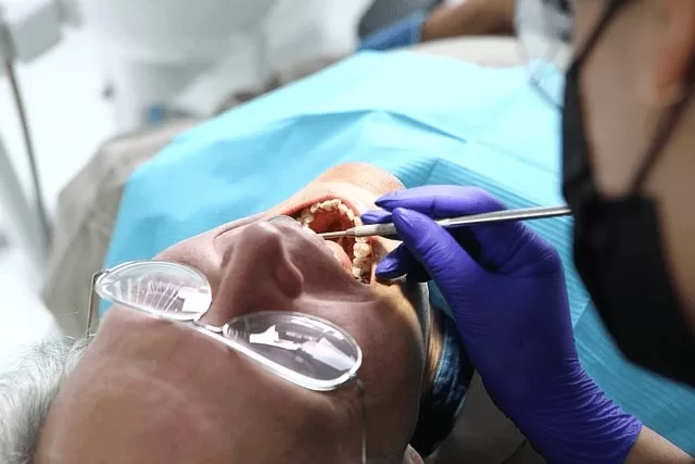 A patient lies back in a dental chair with their mouth open during a procedure, possibly for oral biopsies, while a dental professional in gloves and a mask uses an instrument to examine the patient's mouth.