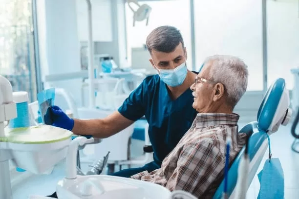 A dentist wearing a mask and gloves shows an older male patient an X-ray image during a consultation, possibly discussing findings related to oral biopsies, inside a modern dental clinic.