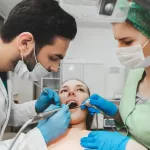 A dentist and assistant wearing masks and gloves performing an oral surgery procedure on a patient in a dental chair. A dentist and assistant wearing masks and gloves performing an oral surgery procedure on a patient in a dental chair.