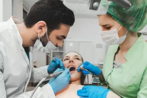 A dentist and assistant wearing masks and gloves performing an oral surgery procedure on a patient in a dental chair. A dentist and assistant wearing masks and gloves performing an oral surgery procedure on a patient in a dental chair.