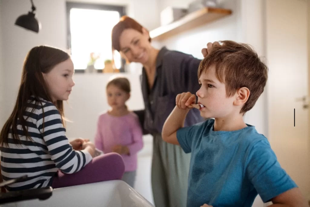 Children practicing oral care in Morristown, with a boy brushing his teeth while supervised by an adult, as other children watch in a family bathroom setting.