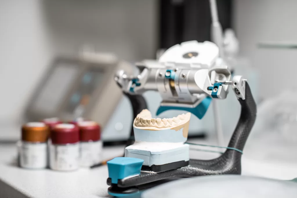 A dental articulator holding a jaw model, used for simulating the movement of a patient's jaw during dental procedures. The setup is placed on a workbench in a dental lab, with jars of dental materials and tools in the background. The precise and modern equipment highlights the meticulous process involved in creating dental restorations.
