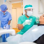 A dentist in green medical clothes examines a patient on a reclining chair. A nurse assists the dentist while setting out the tools for examination.