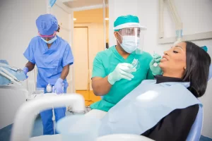 A dentist in green medical clothes examines a patient on a reclining chair. A nurse assists the dentist while setting out the tools for examination.