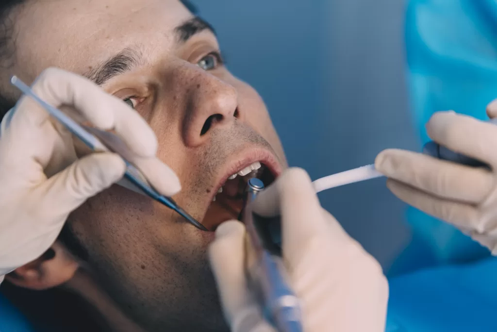 A close-up of a patient undergoing a dental procedure, with gloved hands holding various dental tools, including a mirror and suction device, inside the patient's mouth. The patient looks relaxed as the dentist works, with a clean and professional blue-toned background emphasizing a sterile and clinical environment.