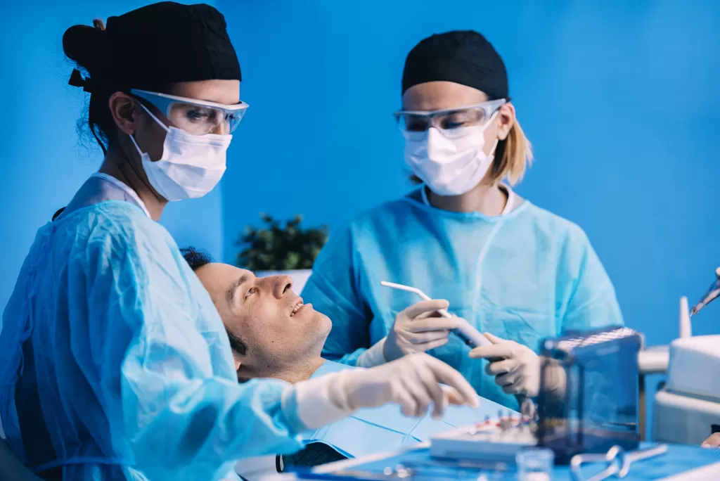 A dentist examines the oral health of a patient through advanced medical equipment. The dentist is assisted by two nurses.