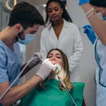 A dentist in a blue uniform and mask performing a dental procedure on a female patient in a dental chair.