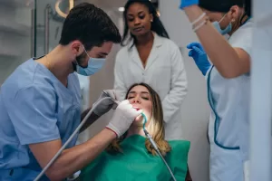 A dentist in a blue uniform and mask performing a dental procedure on a female patient in a dental chair.