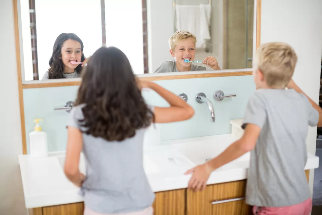 Two children brushing their teeth in front of a bathroom mirror, promoting good oral hygiene habits.