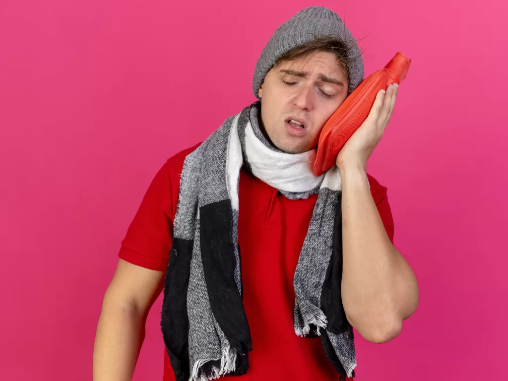 A man wearing a scarf presses a hot water bottle to his cheek, appearing to be in discomfort, likely from pain related to dental surgery.