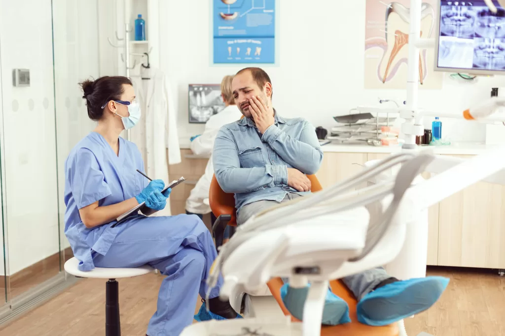 Patient in a dental surgery clinic holding his cheek in pain, while a nurse in blue scrubs examines him and takes notes, showcasing a typical dental surgery consultation scene.