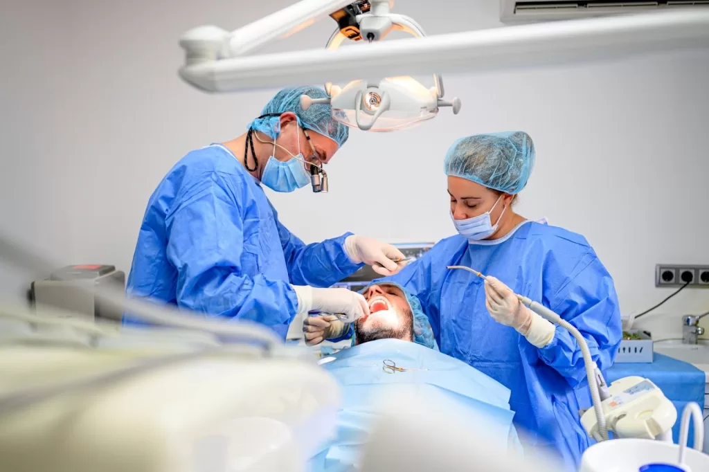 Two dentists in surgical attire perform dental surgery on a patient lying on a chair under a bright surgical light, using specialized tools in a modern dental clinic.