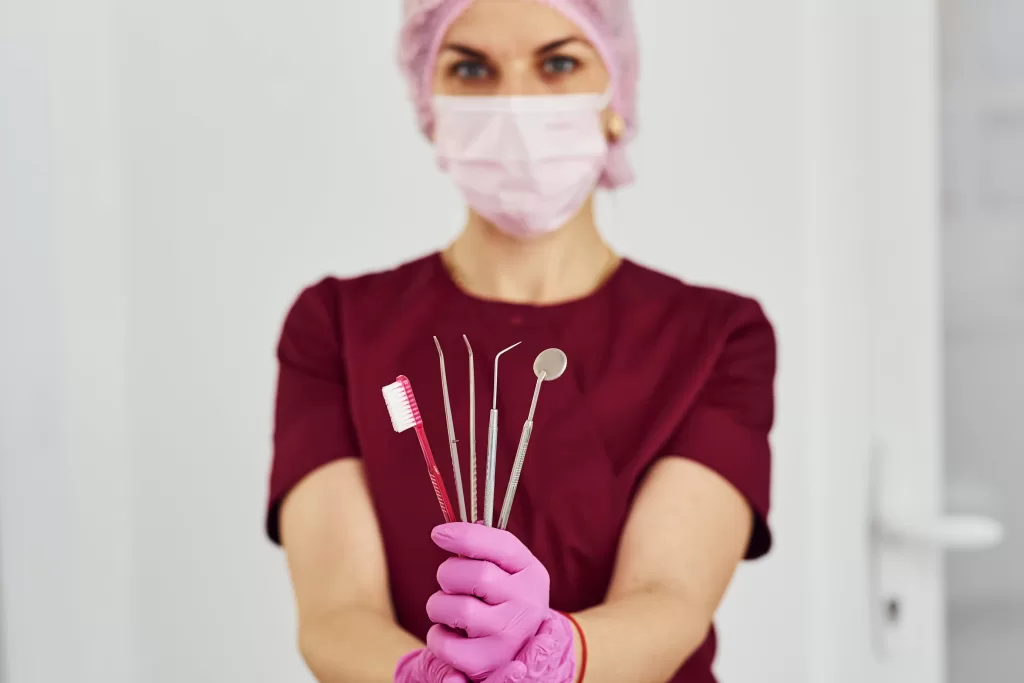 A female dentist wearing a burgundy uniform, pink gloves, and a pink surgical mask holds up various dental tools, including a toothbrush, dental mirror, and probes, in a well-lit clinic.