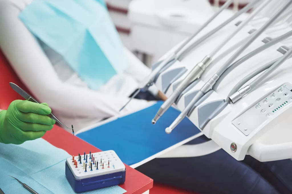 A dentist wearing green gloves holds a dental tool near a tray of precision dental instruments, while a patient reclines in a dental chair covered with a blue protective bib in a modern clinic.