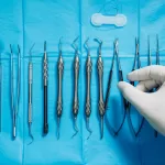 A set of stainless steel dental extraction tools neatly arranged on a blue sterile sheet, with a gloved hand selecting one of the instruments.