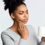 Young woman holding a cup, touching her cheek in discomfort, experiencing tooth sensitivity or dental pain.