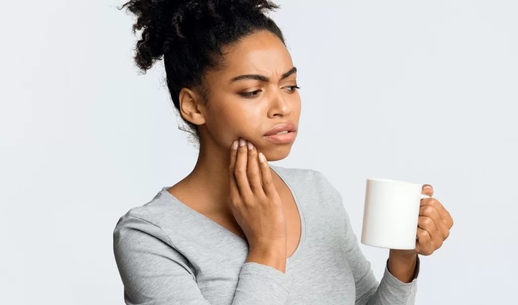 Young woman holding a cup, touching her cheek in discomfort, experiencing tooth sensitivity or dental pain.