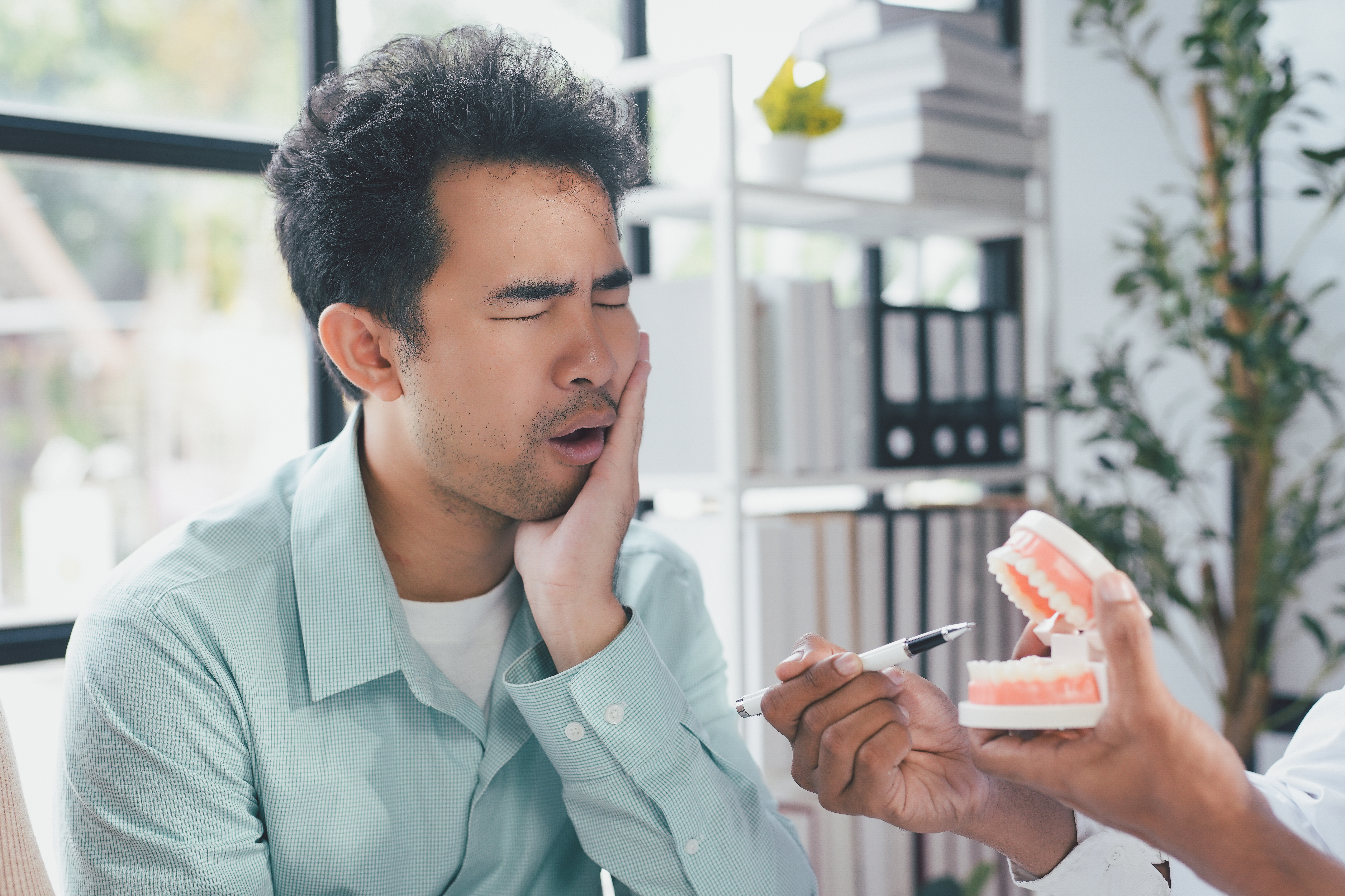 Man holding his jaw in pain while a dentist explains a dental issue using a tooth model inside the Ridge Oral Surgery clinic.