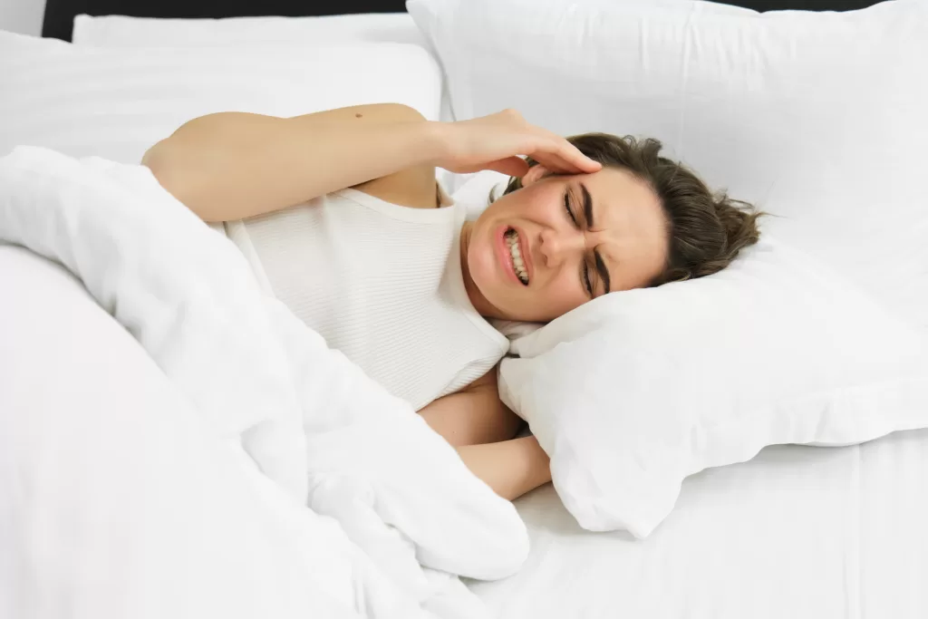 A woman in bed grimacing from a painful headache caused by bruxism.