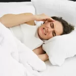 A woman in bed grimacing from a painful headache caused by bruxism.
