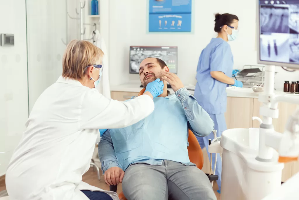 A man in pain at a dental clinic, with a dentist and assistant attending to his jaw issues.