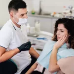 A woman exhibits pain in her jaw while sitting in a dental clinic, with a dentist nearby.