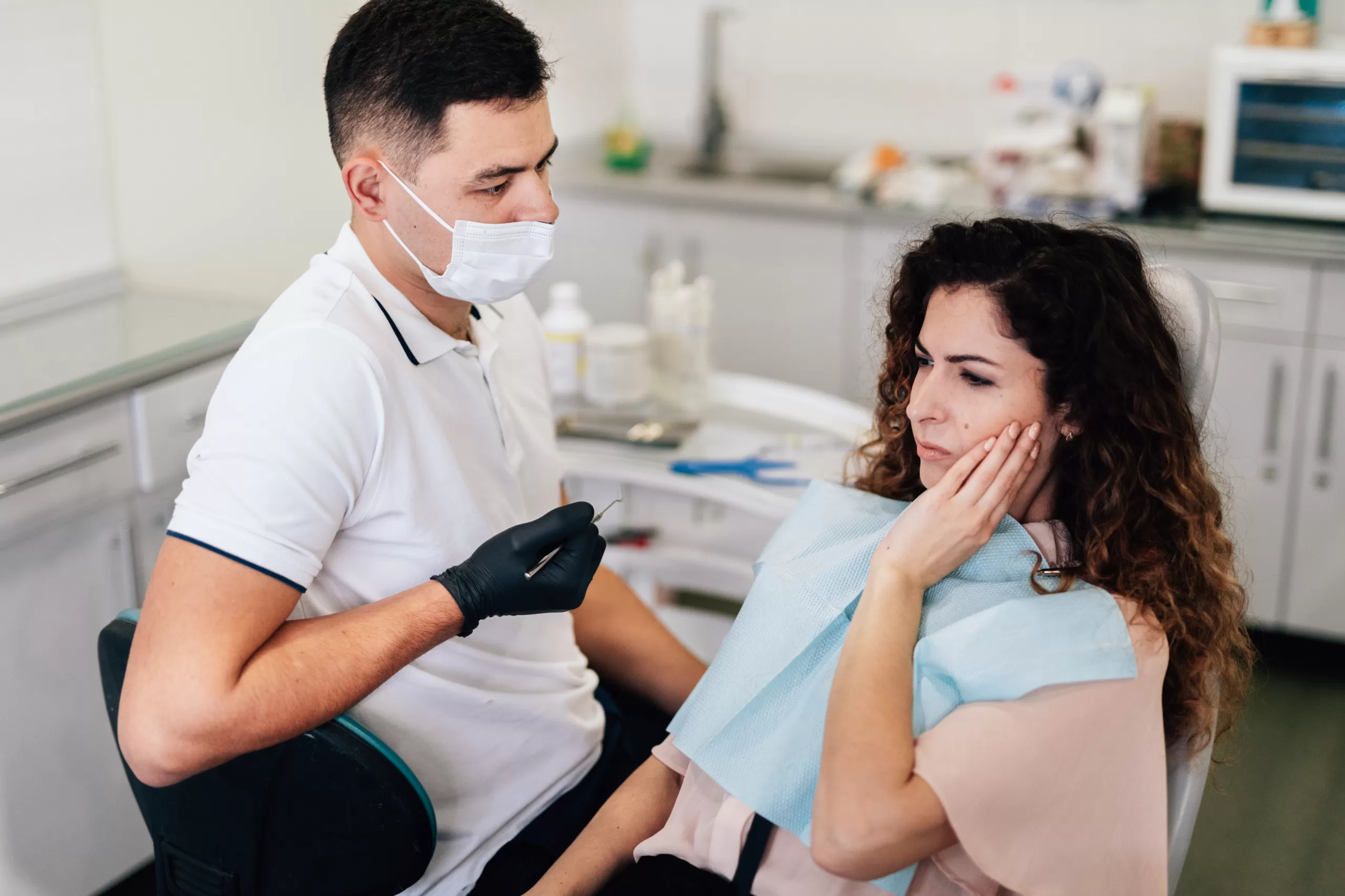 A woman exhibits pain in her jaw while sitting in a dental clinic, with a dentist nearby.