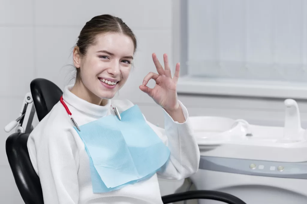 A smiling female patient sitting in a dental chair, wearing a bib, and making an “OK” hand gesture.