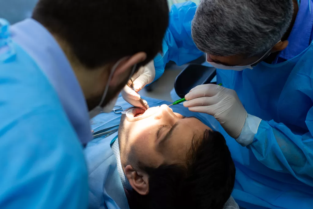 Two dental professionals in blue surgical gowns examine a patient's mouth under bright light during a clinical dental procedure.