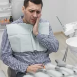 A man in a dental chair holding his jaw in discomfort, surrounded by dental surgery instruments.
