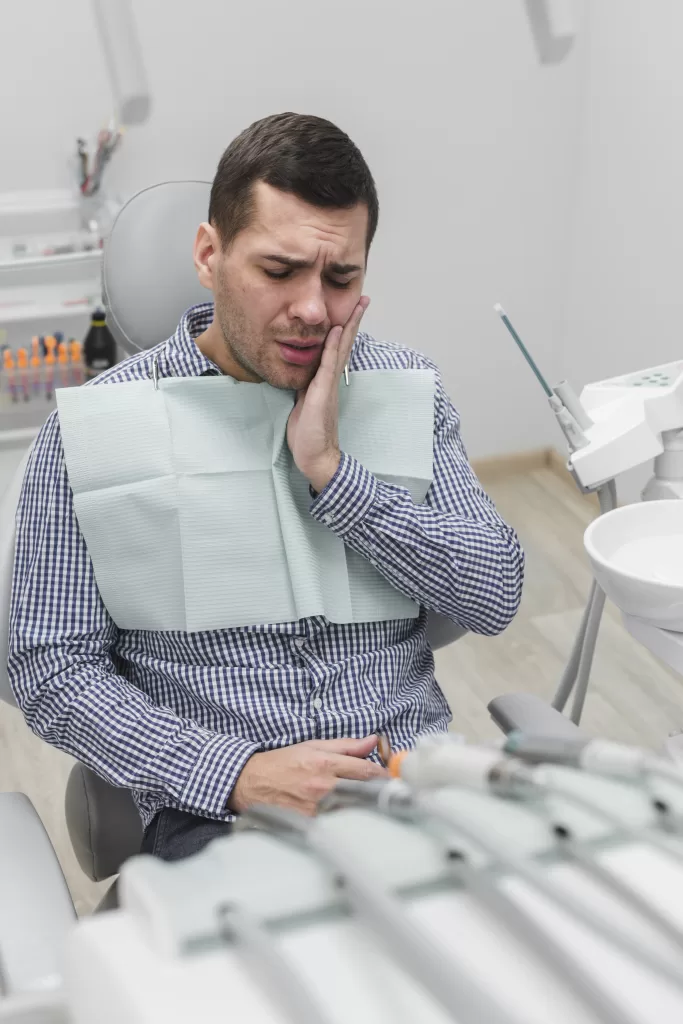 A man in a dental chair holding his jaw in discomfort, surrounded by dental surgery instruments.