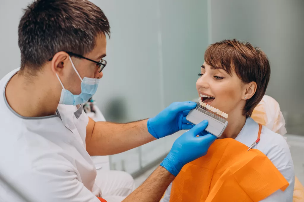 A dentist in scrubs and a mask showing a dental model to a smiling patient sitting in a dental chair.