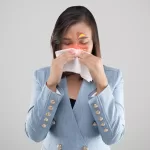 A woman in a blue blazer sneezes into a napkin due to sinus symptoms.