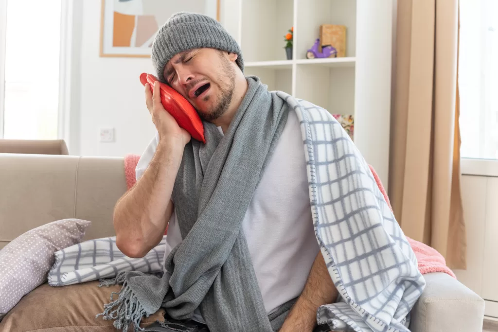 A man wearing a knit cap and shawl, writhing in tooth pain and sitting on a sofa with a hot water bottle.